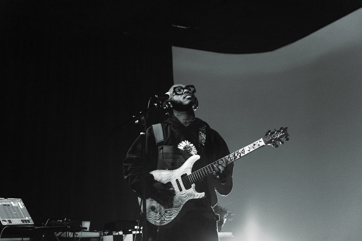 A black and white photograph of a musician on stage, wearing glasses and a dark jacket while playing an electric guitar with a floral graphic on the headstock.